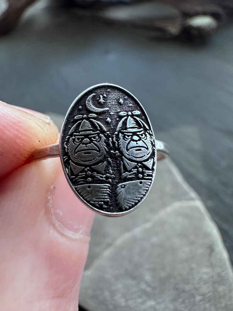 May include: Silver ring with an oval face featuring two stylized figures wearing hats, under a crescent moon and stars. The figures have stern expressions. The ring is held against a neutral background.