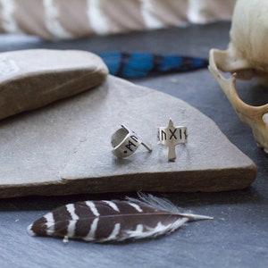 May include: Two silver hoop earrings with a silver arrow design. The arrow has a small inscription of Roman numerals. The earrings are on a gray stone surface.