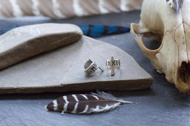 May include: Two silver hoop earrings with a silver arrow design. The arrow has a small inscription of Roman numerals. The earrings are on a gray stone surface.