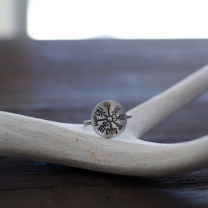 May include: A silver ring with a compass design featuring a circular center with eight points and runes around the edge. The ring is resting on a white antler.