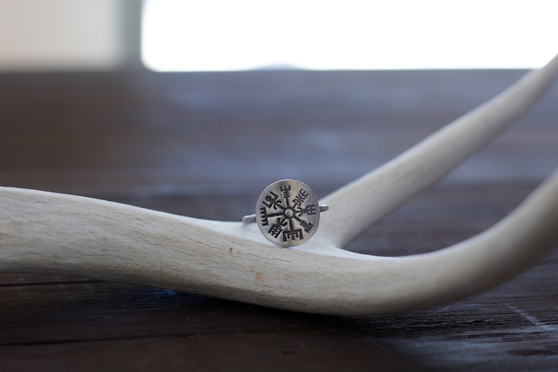 May include: A silver ring with a compass design featuring a circular center with eight points and runes around the edge. The ring is resting on a white antler.