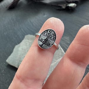 May include: A silver ring with an oval face featuring a detailed engraving of two figures under a crescent moon and stars. The ring is shown on a finger, showcasing the intricate design and craftsmanship of the jewelry.
