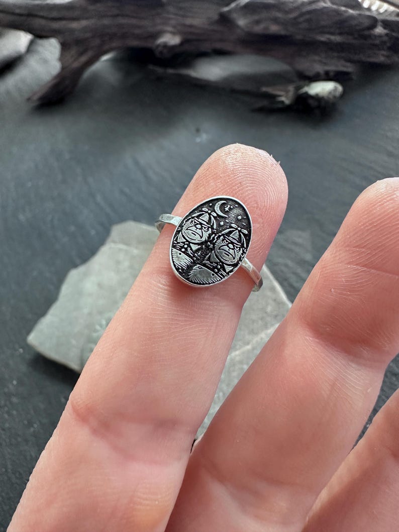 May include: A silver ring with an oval face featuring a detailed engraving of two figures under a crescent moon and stars. The ring is shown on a finger, showcasing the intricate design and craftsmanship of the jewelry.