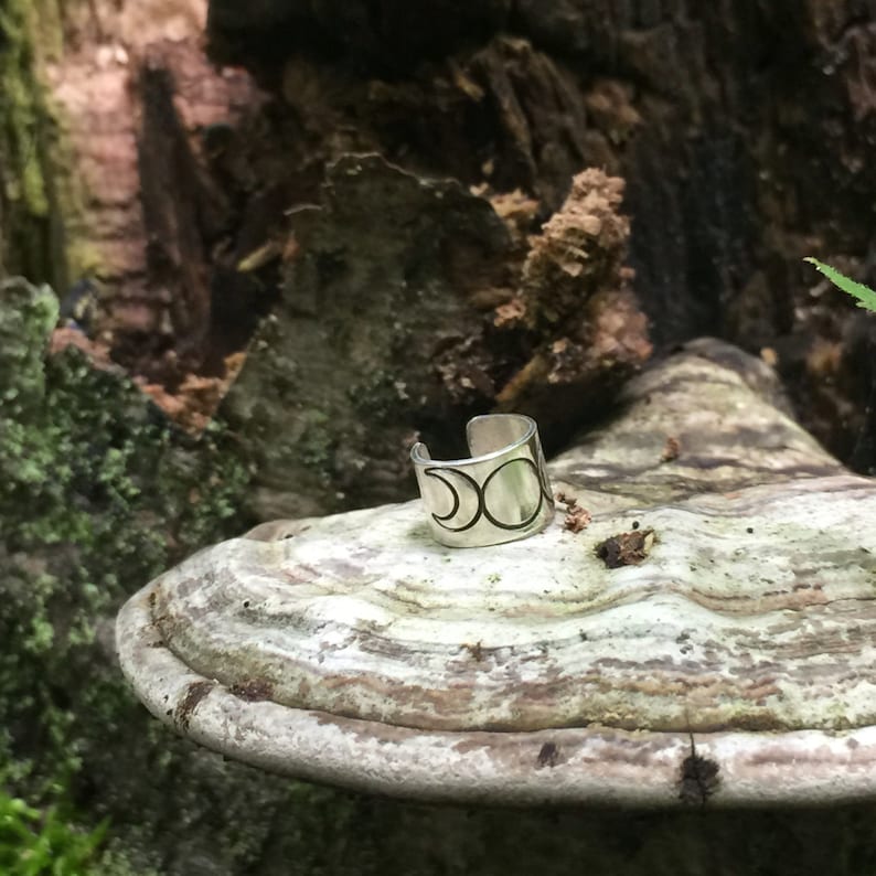 May include: Silver ring with a triple moon design. The ring is on a light brown mushroom-shaped surface.