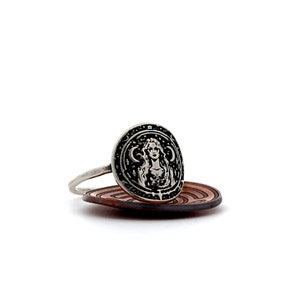 May include: A silver ring with a round face featuring a detailed design of a woman with long hair and a crescent moon on either side of her head. The ring is resting on a brown wooden surface.