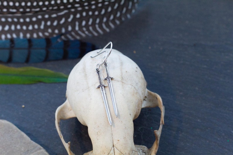 Peut inclure: Une paire de boucles d'oreilles en forme d'&eacute;p&eacute;e argent&eacute;es suspendues &agrave; un cr&acirc;ne blanc. Les &eacute;p&eacute;es sont d&eacute;taill&eacute;es et ont un aspect l&eacute;g&egrave;rement vieilli.