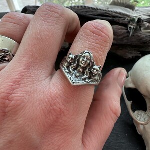 K&ouml;nnte beinhalten: Ein silberner Ring mit einem Frauengesicht, flankiert von zwei Katzen. Der Ring hat ein detailliertes, Vintage-Design mit einer V-f&ouml;rmigen Fassung. Der Ring wird an einem Finger getragen.