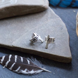 May include: Two silver rings with Roman numeral engravings. The rings are on a gray stone surface. The rings are in the shape of a bird with its wings spread.