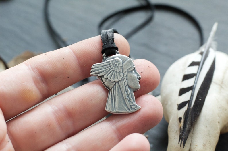 May include: Silver pendant necklace featuring a profile of a woman with a winged headdress. The pendant is on a black leather cord.