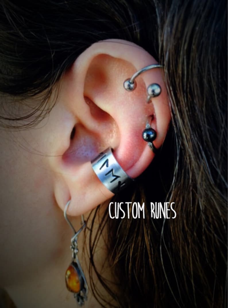 May include: A close-up of a person's ear with multiple piercings. The ear is adorned with a silver ear cuff with custom runes, a silver hoop earring with a black bead, and a silver barbell with two silver beads. The text "CUSTOM RUNES" is visible below the ear cuff.