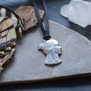 May include: A silver pendant necklace featuring a profile of a woman with a feathered headdress. The necklace is on a black leather cord and is displayed on a gray stone surface.