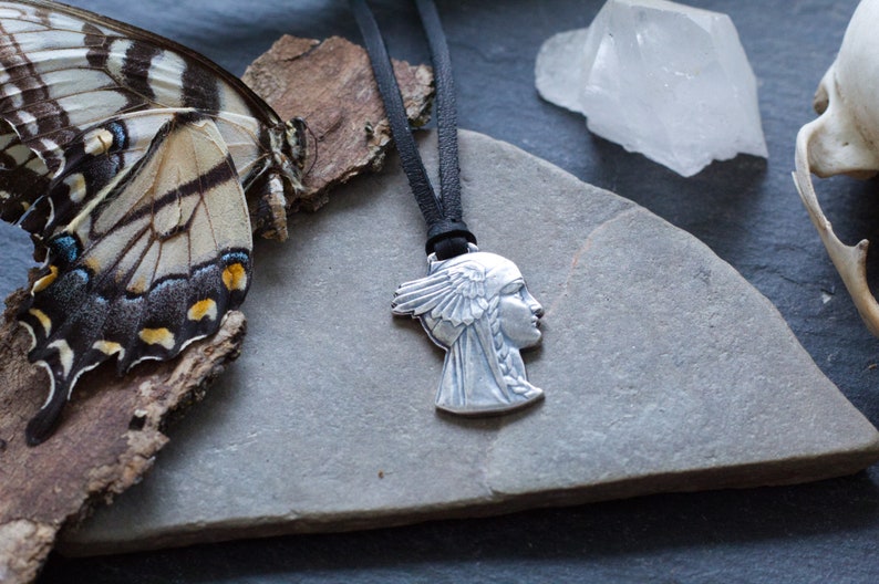 May include: A silver pendant necklace featuring a profile of a woman with a feathered headdress. The necklace is on a black leather cord and is displayed on a gray stone surface.