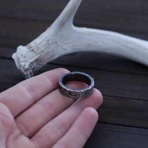 May include: A silver ring with an intricate design, held in a hand against a dark wooden background. A white antler is visible in the background.