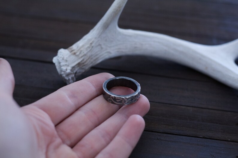May include: A silver ring with an intricate design, held in a hand against a dark wooden background. A white antler is visible in the background.