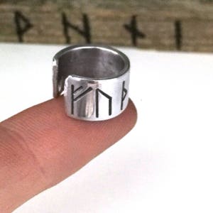 May include: A silver ring with a band that is engraved with four runes. The ring is open on one side and is worn on a finger.