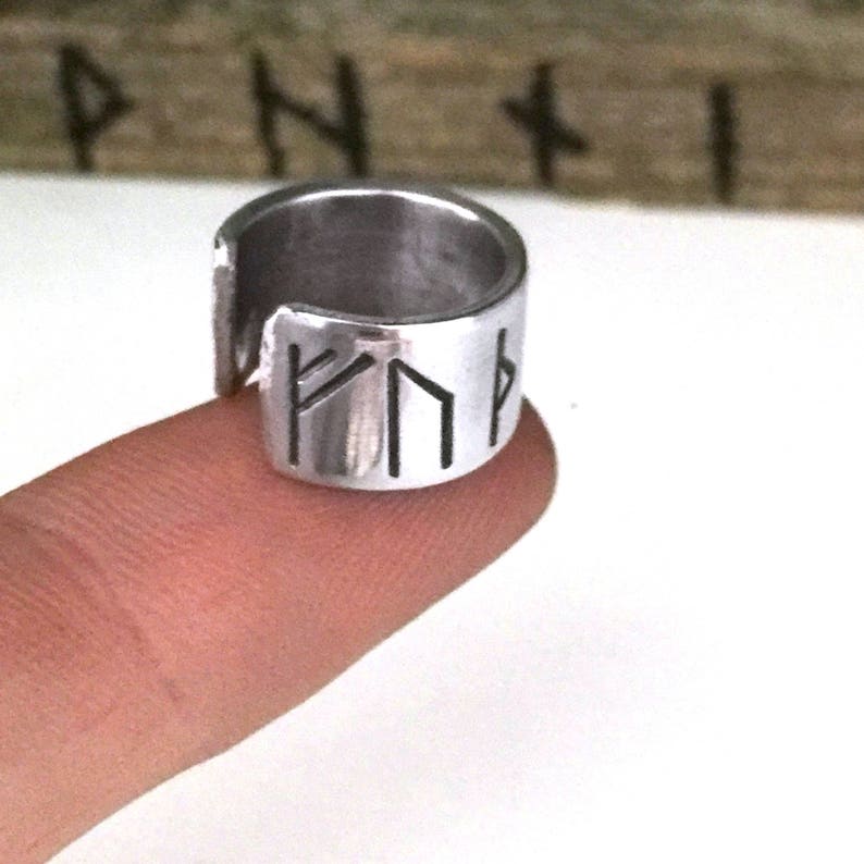May include: A silver ring with a band that is engraved with four runes. The ring is open on one side and is worn on a finger.