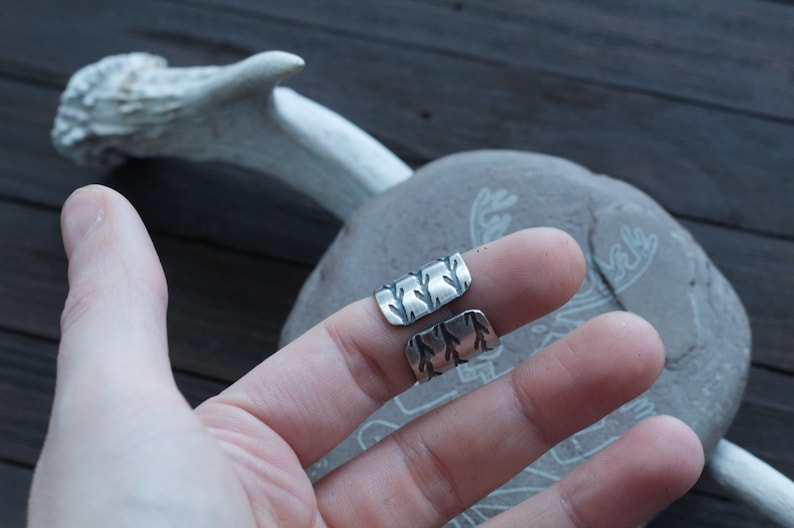 May include: Two silver rings with a textured design. The rings are worn on the index and middle fingers of a hand.