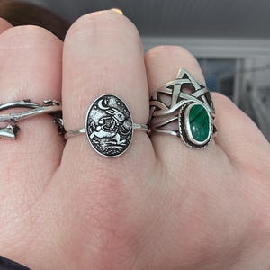 May include: A hand displays three silver rings. One ring features an oval design with a rabbit and moon scene. Another ring has a green oval stone set in a Celtic knot design. The third ring has a vine-like design.