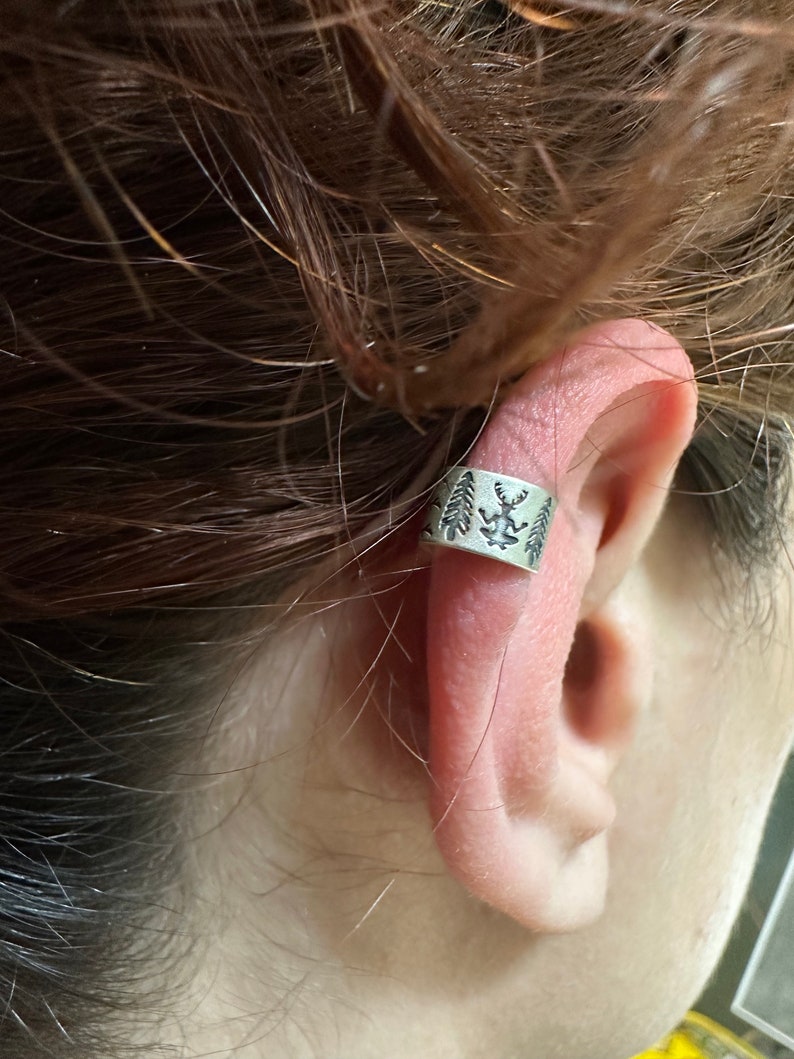 May include: A silver ear cuff with a design of trees and a deer. The cuff is worn on the upper part of the ear.