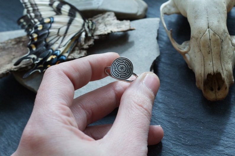 May include: A silver ring featuring a circular design with a maze-like pattern. The ring is held in a hand, with a butterfly and a skull in the background. The ring's design is detailed and intricate.
