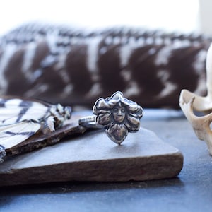 May include: A silver ring with a detailed floral design featuring a face in the centre. The ring is resting on a piece of wood with a butterfly wing in the background.