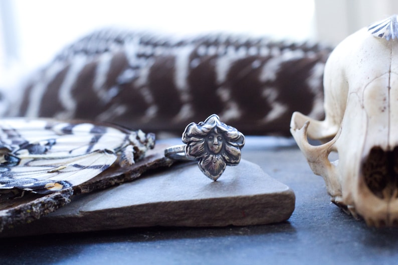 May include: A silver ring with a detailed floral design featuring a face in the centre. The ring is resting on a piece of wood with a butterfly wing in the background.