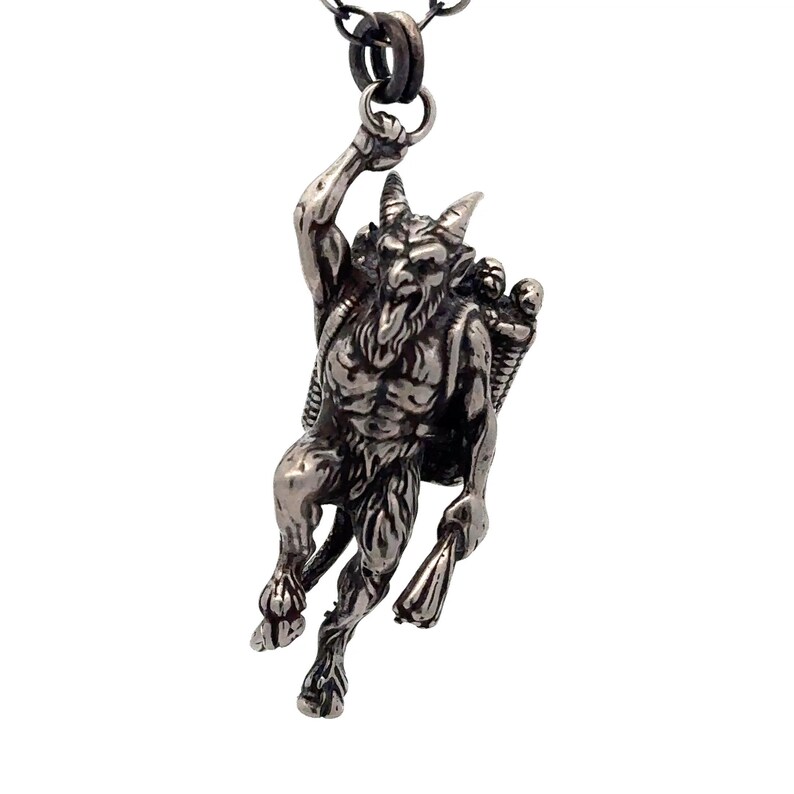 May include: Silver-toned pendant necklace featuring a detailed depiction of a horned, goat-legged figure carrying a basket and a torch. The pendant is suspended from a delicate chain, showcasing a gothic or mythological theme.