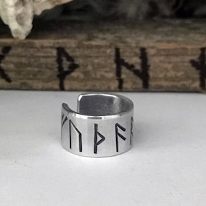 May include: A silver ring with a band of runes engraved on it. The runes are black and the ring is made of metal.