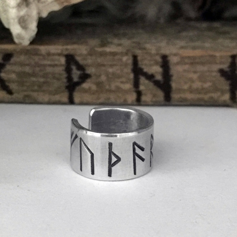 May include: A silver ring with a band of runes engraved on it. The runes are black and the ring is made of metal.