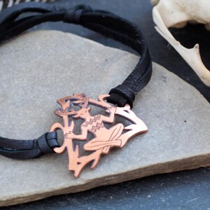 May include: A black leather bracelet with a copper pendant featuring a stylized figure with antlers. The pendant is intricately detailed and has a rustic, earthy aesthetic.