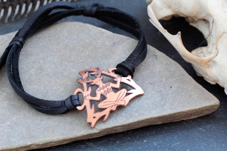 May include: A black leather bracelet with a copper pendant featuring a stylized figure with antlers. The pendant is intricately detailed and has a rustic, earthy aesthetic.