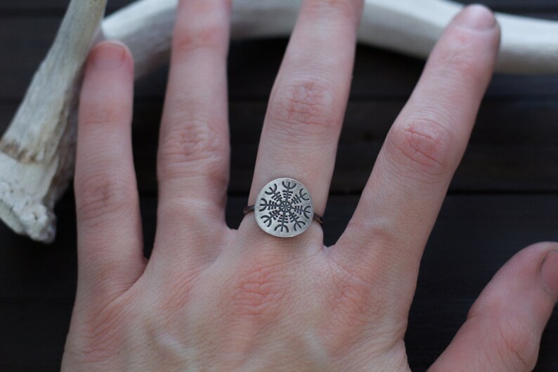 May include: A silver ring with a circular design featuring a Vegvisir symbol, a Norse directional symbol, on a person's finger.