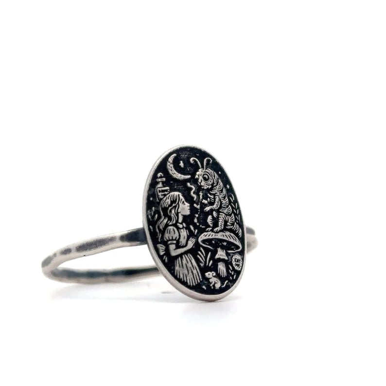 May include: Silver-toned ring featuring an oval pendant with an engraved scene from Alice in Wonderland. The design includes Alice, a caterpillar, mushrooms, and a crescent moon. The ring has a textured band.