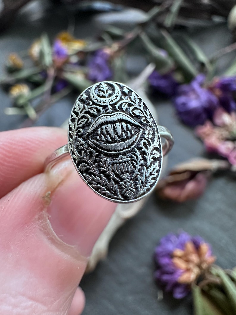 May include: A silver ring with an oval face featuring a detailed design of a mouth with sharp teeth, surrounded by floral and leaf patterns. The ring is held by a person's finger, with a blurred background of purple flowers and greenery.