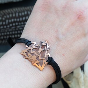 May include: A copper bracelet with a detailed cutout design of a mythical creature. The bracelet is made of black leather and has a simple adjustable closure.
