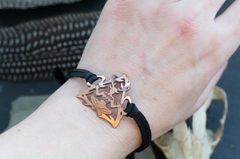 May include: A copper bracelet with a detailed cutout design of a mythical creature. The bracelet is made of black leather and has a simple adjustable closure.