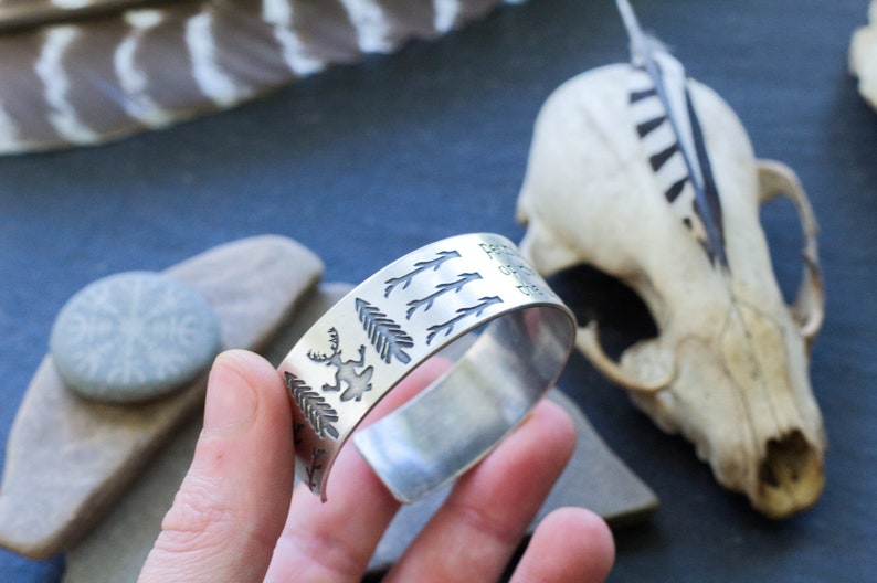 May include: Silver cuff bracelet with an engraved design of trees, deer, and the words "Forever Wild".