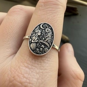 May include: A silver ring featuring an oval pendant with an engraved scene from Alice in Wonderland. The design includes Alice, a caterpillar, a mushroom, and a crescent moon. The ring is worn on a finger.