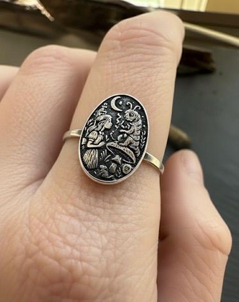 May include: A silver ring featuring an oval pendant with an engraved scene from Alice in Wonderland. The design includes Alice, a caterpillar, a mushroom, and a crescent moon. The ring is worn on a finger.