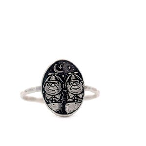 May include: A silver ring featuring an oval-shaped face with two cartoon characters wearing hats and suits, set against a starry night sky with a crescent moon. The ring has a textured band.