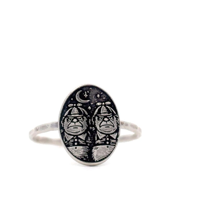 May include: A silver ring featuring an oval-shaped face with two cartoon characters wearing hats and suits, set against a starry night sky with a crescent moon. The ring has a textured band.