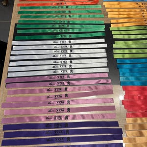 Peut inclure: Une collection de rubans colorés dans les tons de rouge, orange, vert, blanc, rose, violet, bleu et or. Chaque ruban porte l'inscription "XCFA". Les rubans sont disposés avec soin sur une surface en bois.