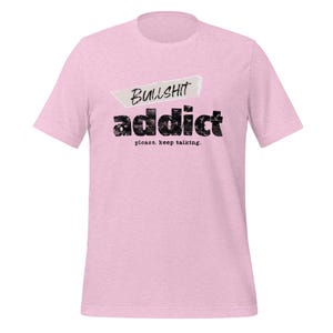 Puede incluir: Camiseta rosa claro con las palabras "BULLSHIT addict" en negro, con una fuente desgastada. La frase "please. keep talking." está debajo de la palabra "addict". La palabra "BULLSHIT" está encima de la palabra "addict" en una forma de cinta blanca.
