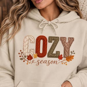 May include: A beige hooded sweatshirt featuring the word COZY embroidered in large, textured letters with a cable-knit pattern in cream, rust orange, and olive green. Below, the word season is stitched in brown, surrounded by autumn leaves.