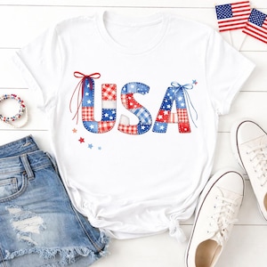 May include: White t-shirt with the word "USA" in a patchwork design of red, white, and blue fabrics. The letters are decorated with bows and stars. The image also shows denim shorts, white sneakers, and small American flags.