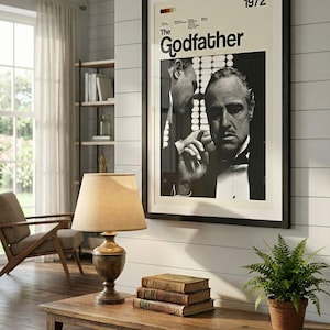 K&ouml;nnte beinhalten: Gerahmtes Schwarz-Wei&szlig;-Poster des Films "Der Pate" von 1972, das an einer wei&szlig;en Wand h&auml;ngt. Ein Holztisch mit B&uuml;chern und einer Lampe steht unter dem Poster. Eine kleine Topfpflanze steht auf dem Tisch.