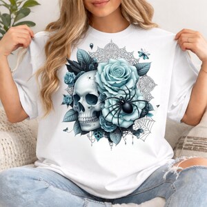 Puede incluir: Una camiseta blanca con un estampado gr&aacute;fico de un cr&aacute;neo humano rodeado de rosas azul claro, hojas oscuras, una ara&ntilde;a negra y un delicado patr&oacute;n de telara&ntilde;a. El dise&ntilde;o incluye peque&ntilde;as mariposas y detalles intrincados tipo encaje.