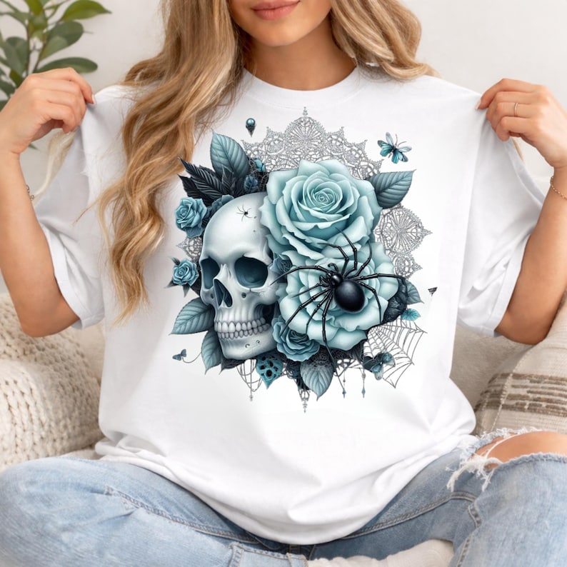 Puede incluir: Una camiseta blanca con un estampado gr&aacute;fico de un cr&aacute;neo humano rodeado de rosas azul claro, hojas oscuras, una ara&ntilde;a negra y un delicado patr&oacute;n de telara&ntilde;a. El dise&ntilde;o incluye peque&ntilde;as mariposas y detalles intrincados tipo encaje.