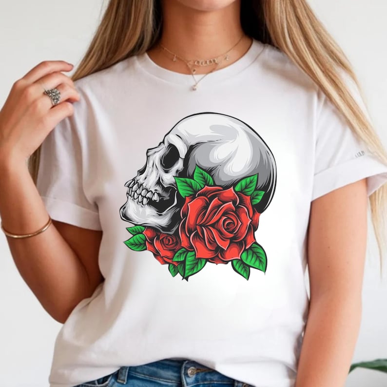 Puede incluir: Una camiseta blanca de manga corta con un estampado gr&aacute;fico de un cr&aacute;neo humano de perfil, situado sobre un grupo de rosas rojas vibrantes con hojas verdes detalladas. El cr&aacute;neo est&aacute; representado en tonos grises y blancos con sombreado.