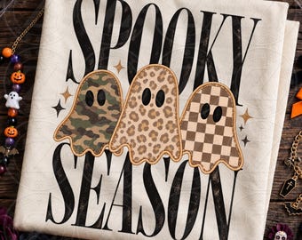 May include: A cream-colored t-shirt featuring the black text SPOOKY SEASON. Between the words are three ghost shapes with stitched edges: one with a green camouflage pattern, one with a tan leopard print, and one with a brown checkerboard pattern.